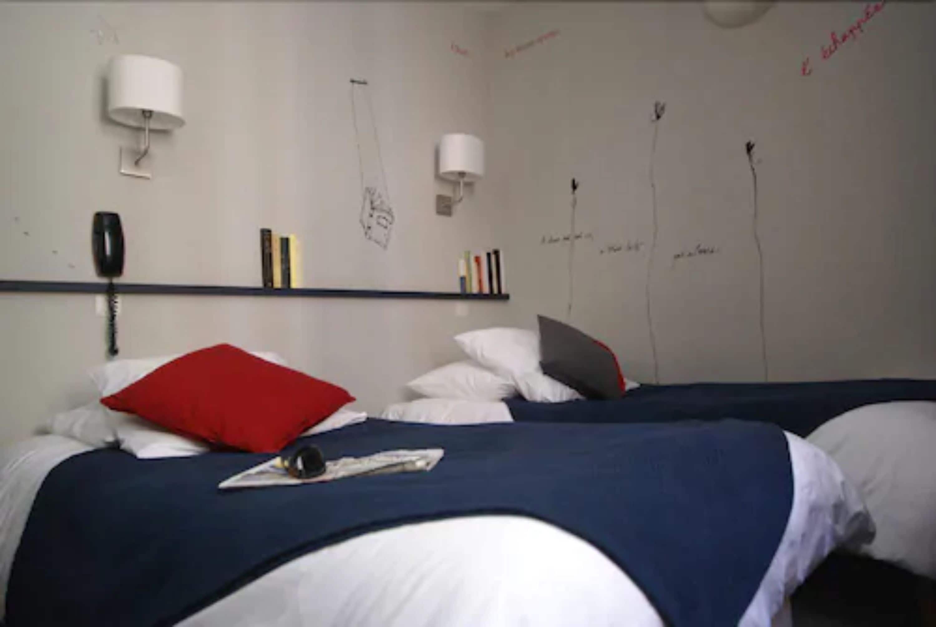 Une chambre d’hôtel avec deux lits, chacun recouvert d’une couette bleue et blanche, agrémenté d’oreillers rouges et blancs, et dotée d’une table de chevet avec une lampe. On y trouve également des livres et une petite étagère fixée au mur.