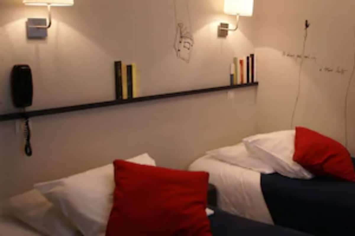 A room with two beds, each with a pillow and a blanket. There is a shelf with books and a telephone on the wall.