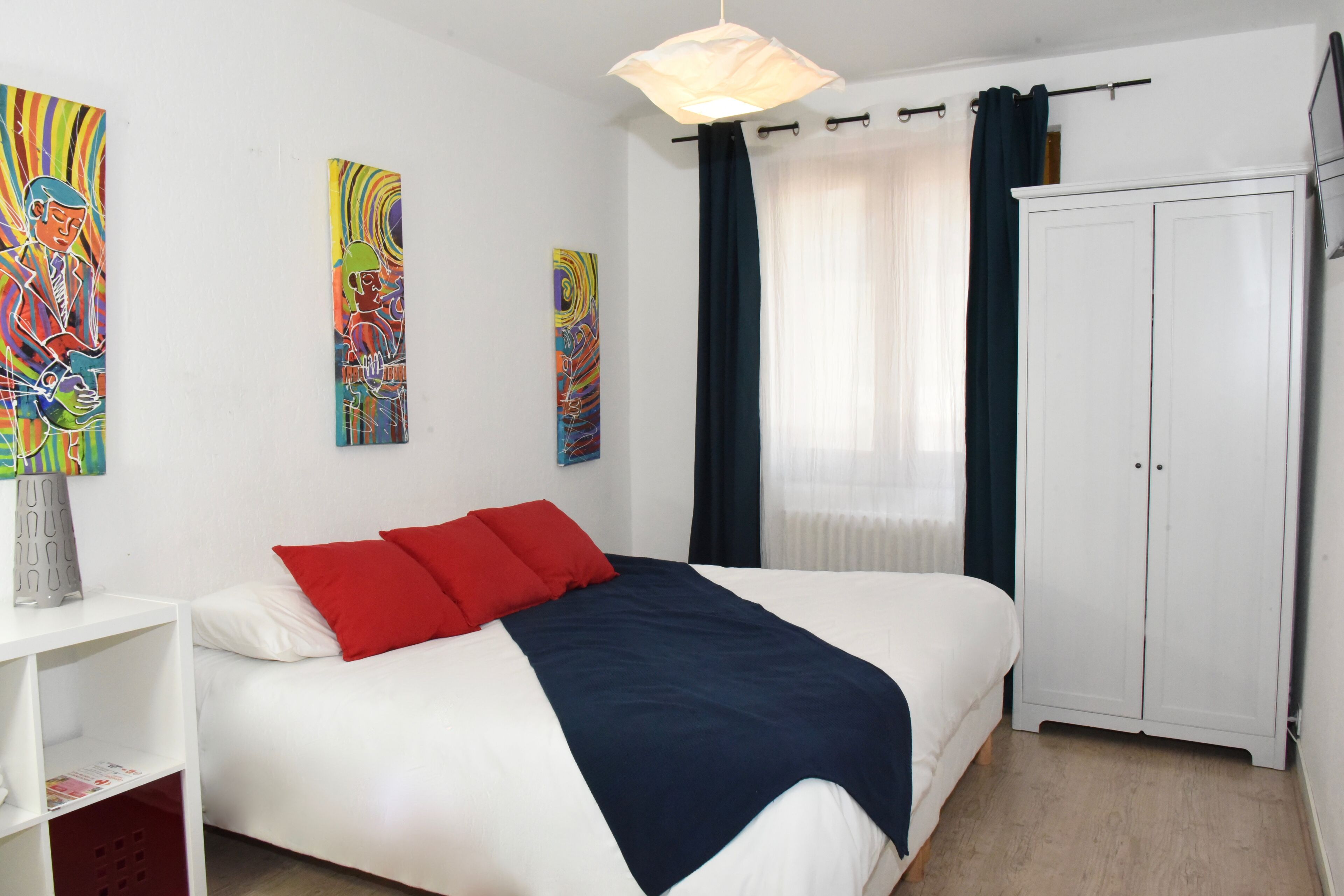 A bedroom with a bed, white wardrobe, and two paintings on the wall.