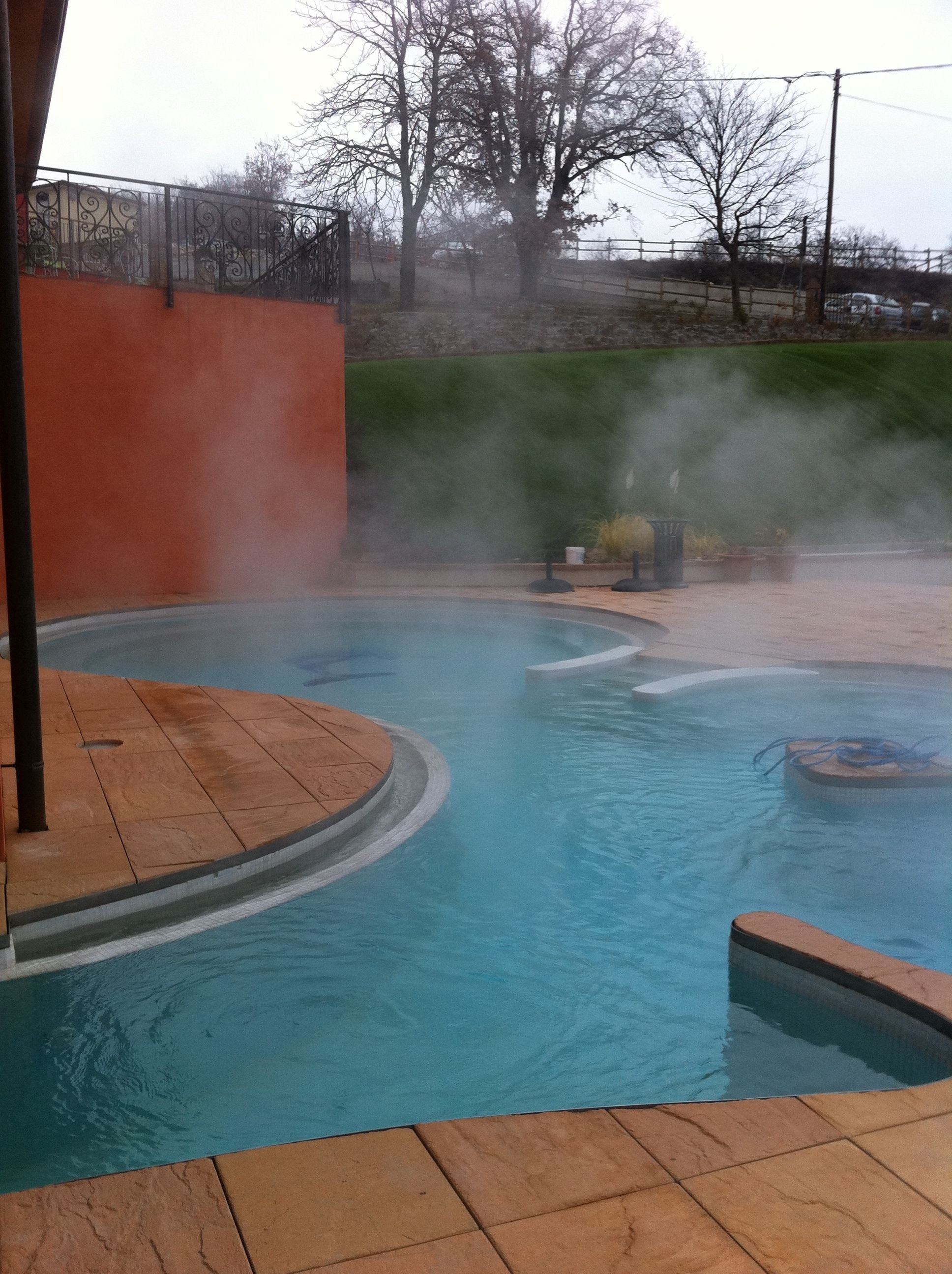 Une piscine d’eau chaude où s’échappe de la vapeur, entourée d’une terrasse en bois et d’un mur de pierre.
