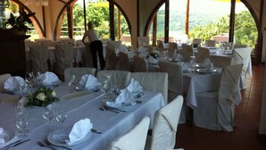 Un salón de banquetes con mesas redondas dispuestas para cenar, amplias ventanas en arco y una araña de cristal.