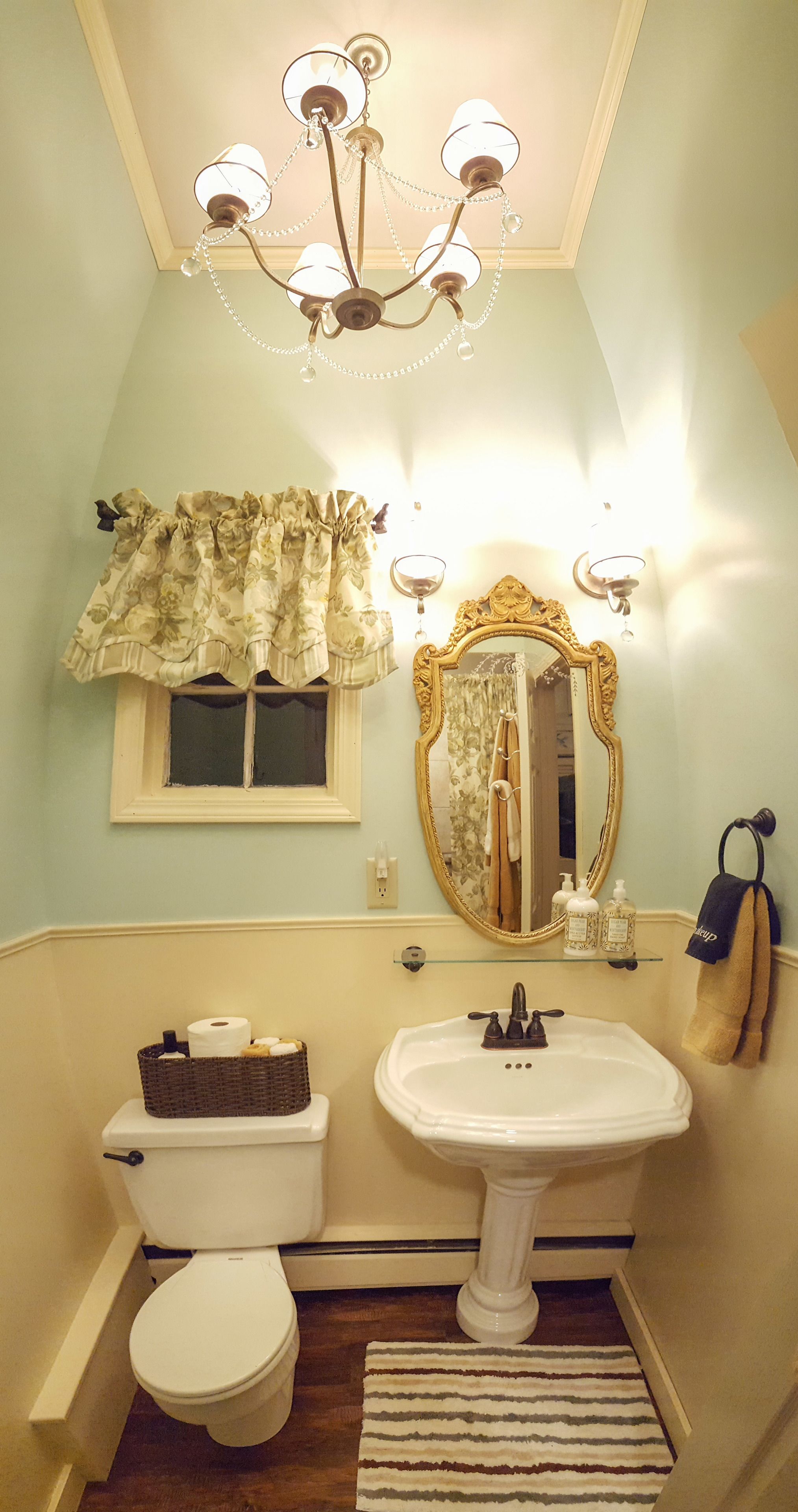 Un baño con inodoro blanco, lavamanos pedestal, espejo con marco decorativo, candelabro y ventana con cortinas florales.