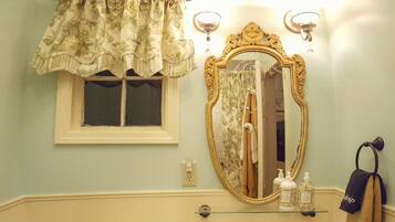 Un baño con inodoro blanco, lavamanos pedestal, espejo con marco decorativo, candelabro y ventana con cortinas florales.