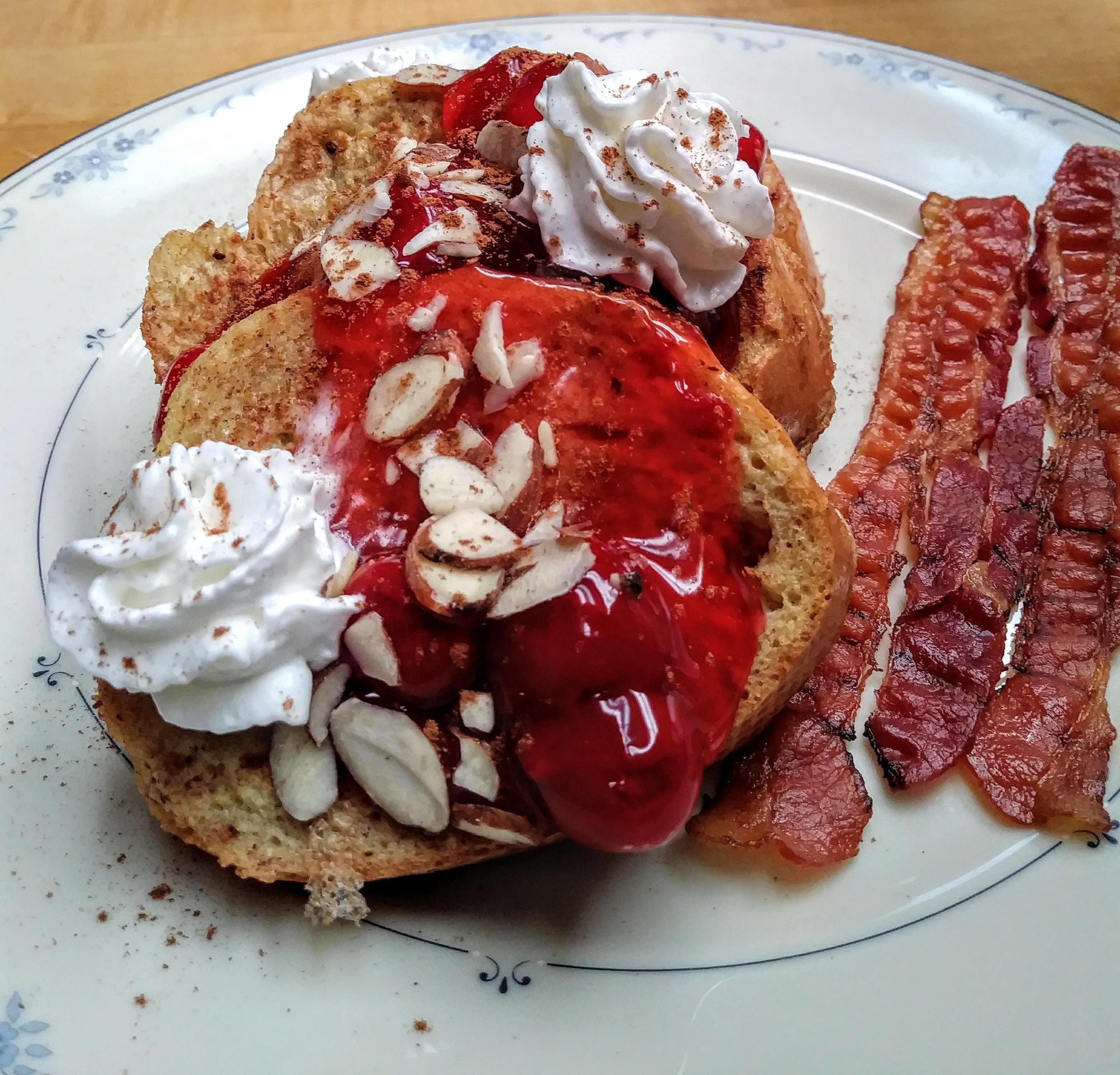 A plate with a breakfast of toast topped with whipped cream, red jam, and sliced almonds, accompanied by a strip of bacon.