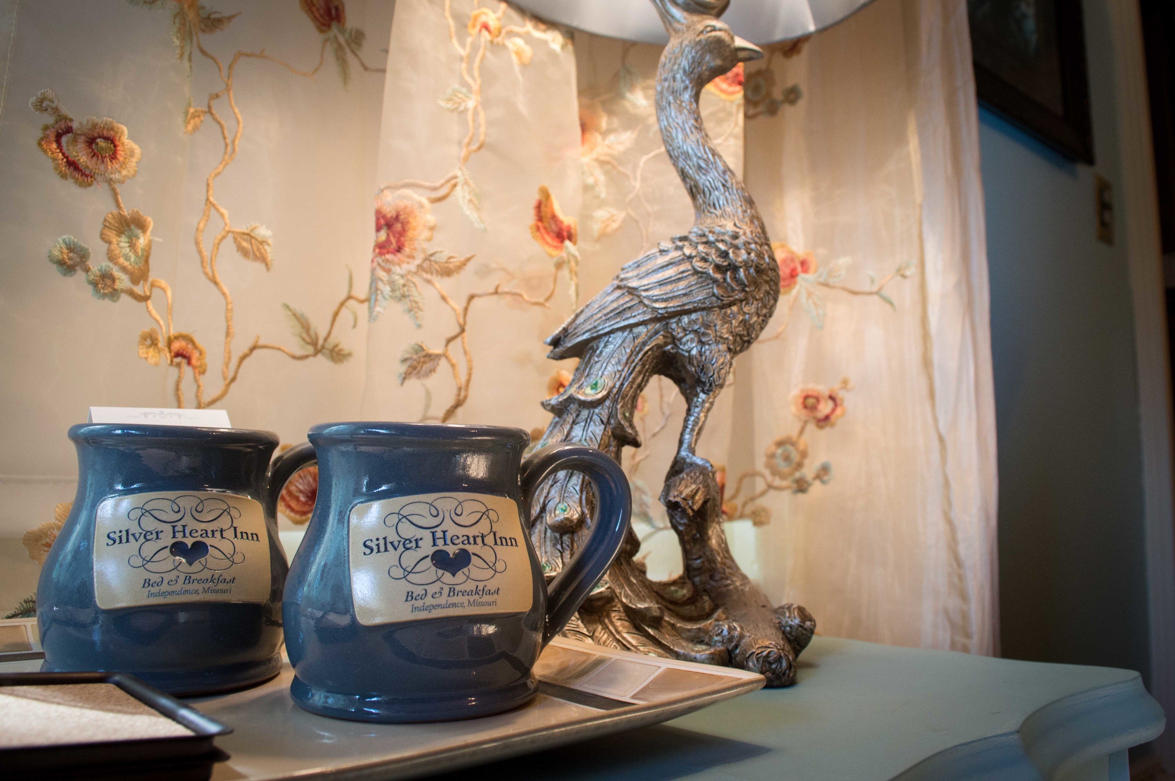 Deux tasses en céramique ornées de l’inscription « Silver Heart Inn Bed & Breakfast, Independence, Missouri », posées sur une table avec une sculpture d’oiseau décorative.