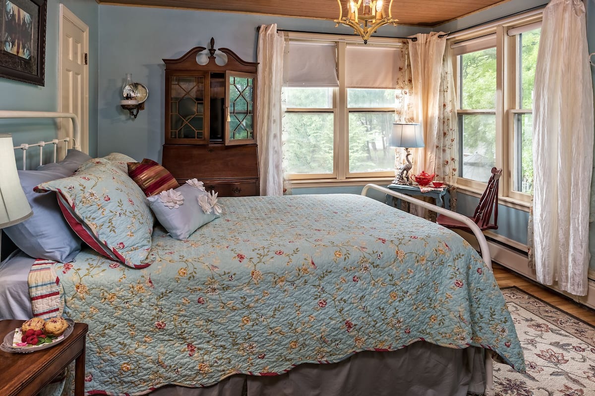 A bedroom with a floral quilted bedspread, a wooden dresser, a bedside table with a lamp, and a window with curtains.
