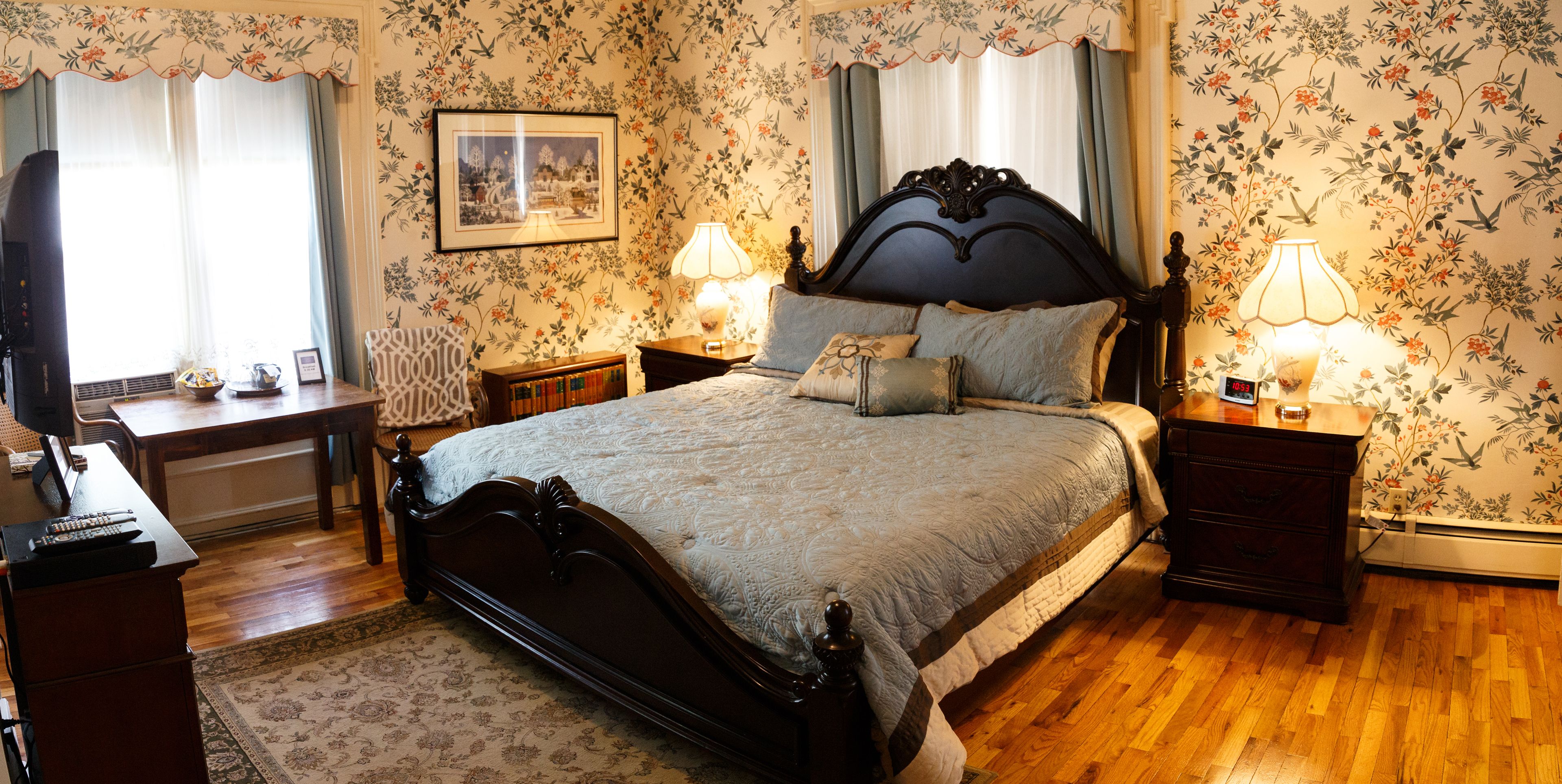 A bedroom with a large bed, two bedside tables with lamps, a desk with a chair, a television, and a framed picture on the wall.