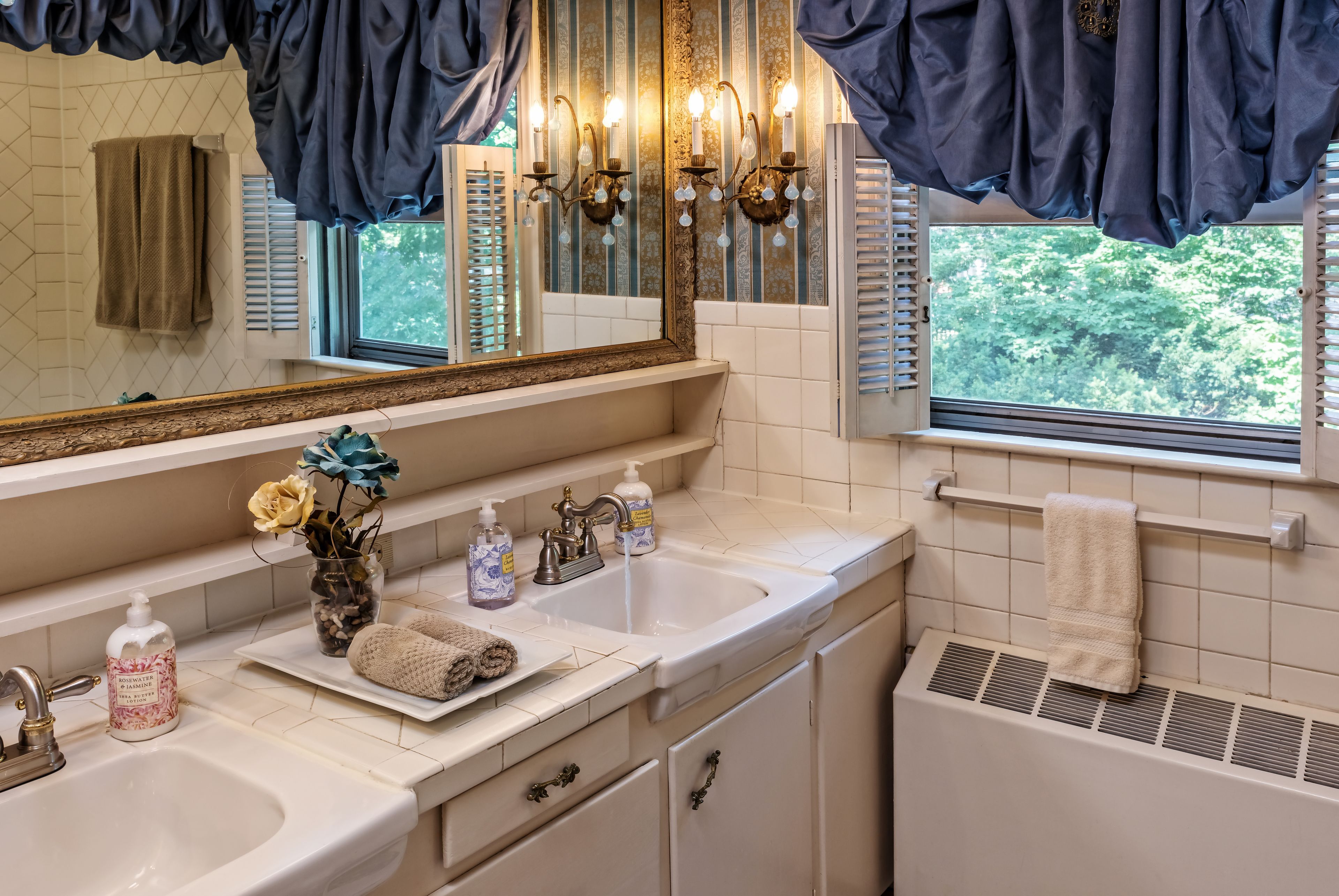 A bathroom with two sinks, a mirror, a window with shutters, and a towel rack.