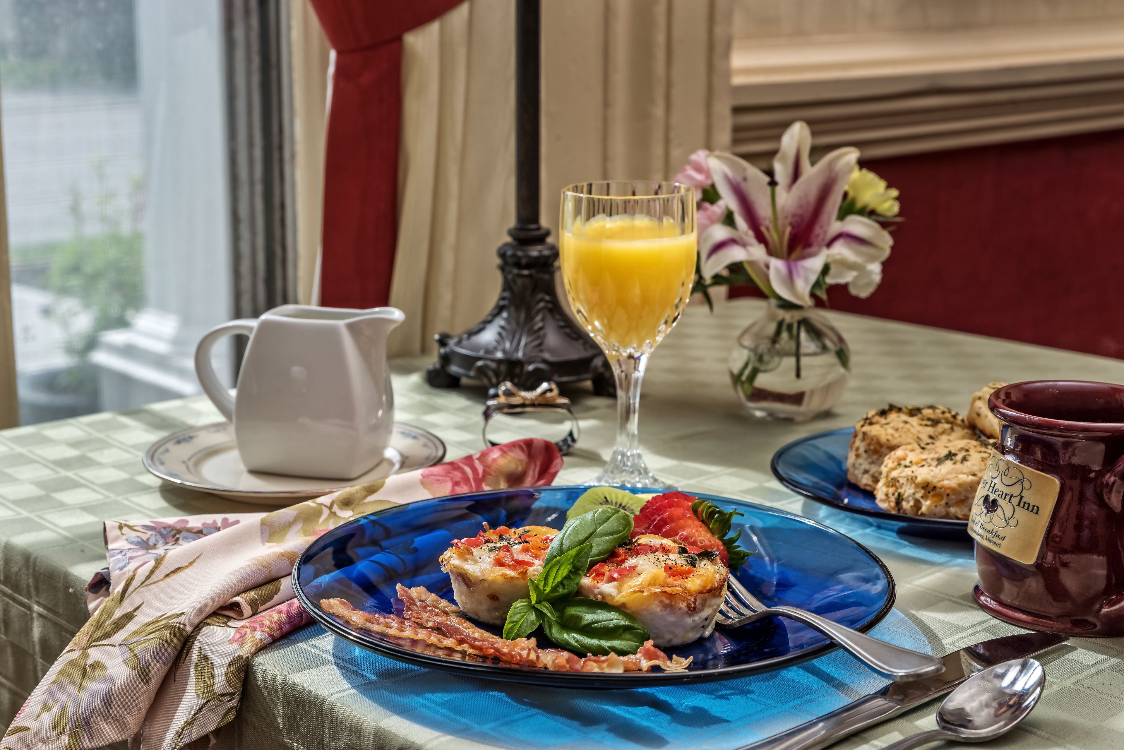 Uma mesa de café da manhã com toalha quadriculada, um prato azul com comida, um copo de suco de laranja e uma caneca vermelha.