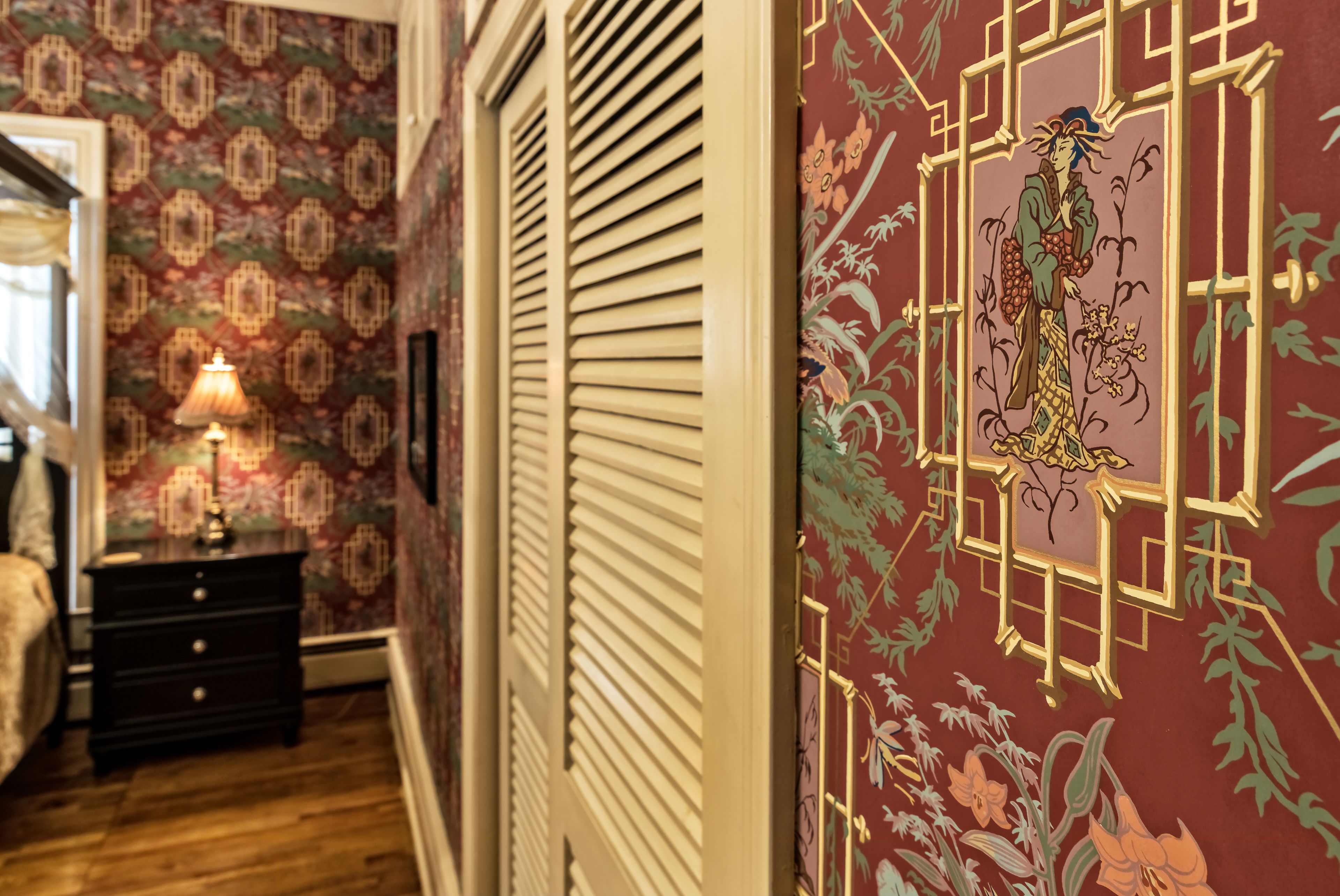 A hallway with patterned wallpaper, a wooden floor, a nightstand with a lamp, and a decorative wall panel.
