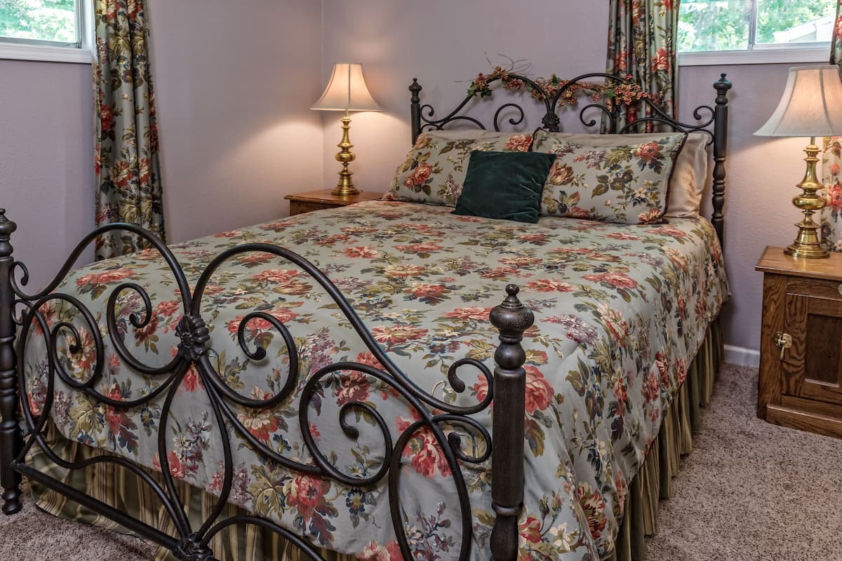 A bedroom with a floral bedspread, a wooden headboard, two bedside tables with lamps, and curtains.
