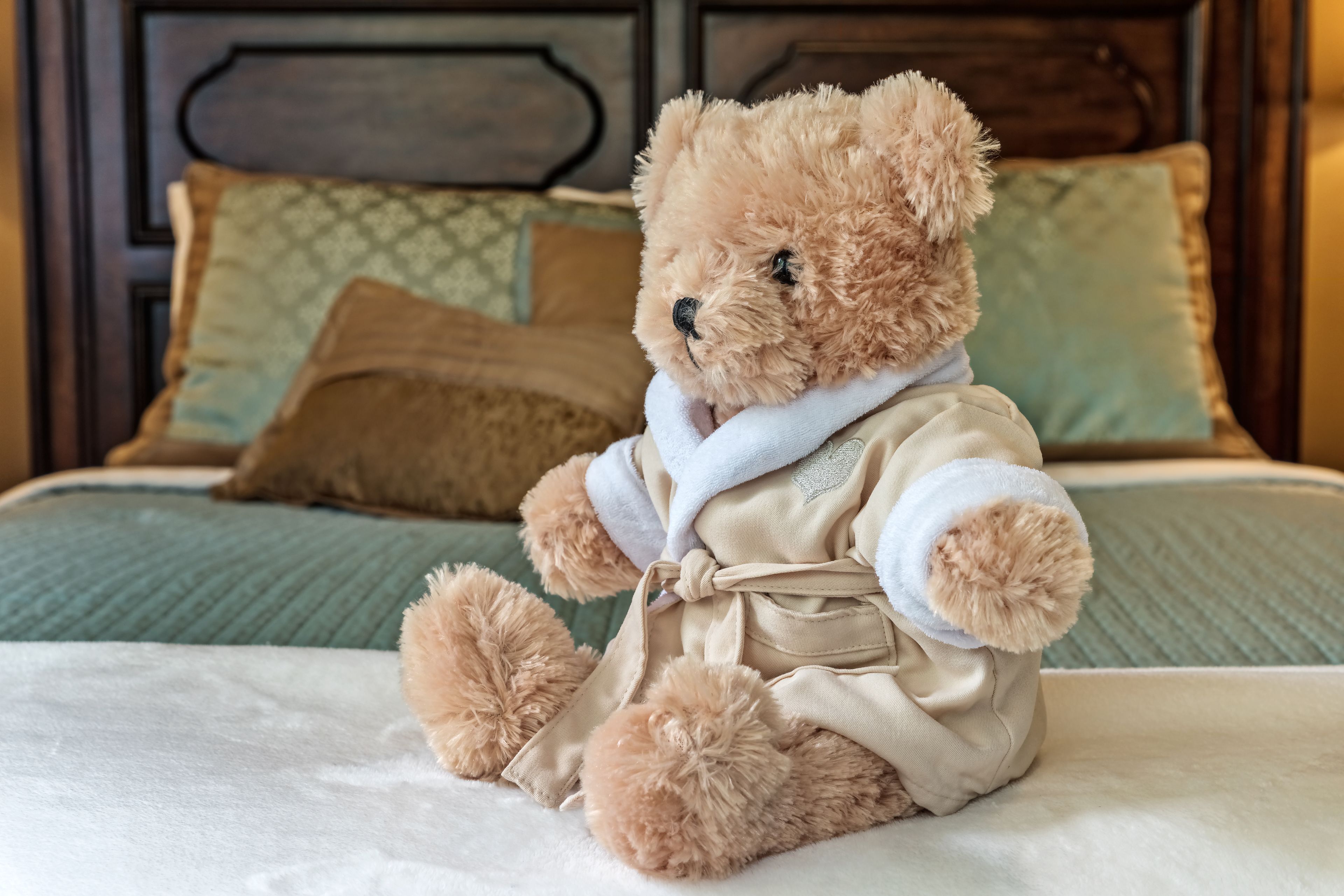 Een teddybeer in een badjas zittend op een bed met een houten hoofdeinde en bedkussens met een patroon.