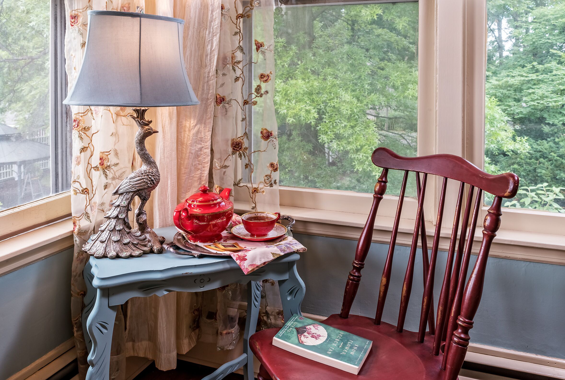 A room with a window, a table with a lamp, a red teapot, a cup, and a chair with a book.