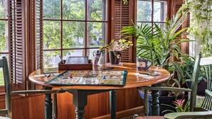 A wooden table with a board game, surrounded by wicker chairs, in a room with wooden shutters and potted plants. - Silver Heart Inn and Cottages (Independence)