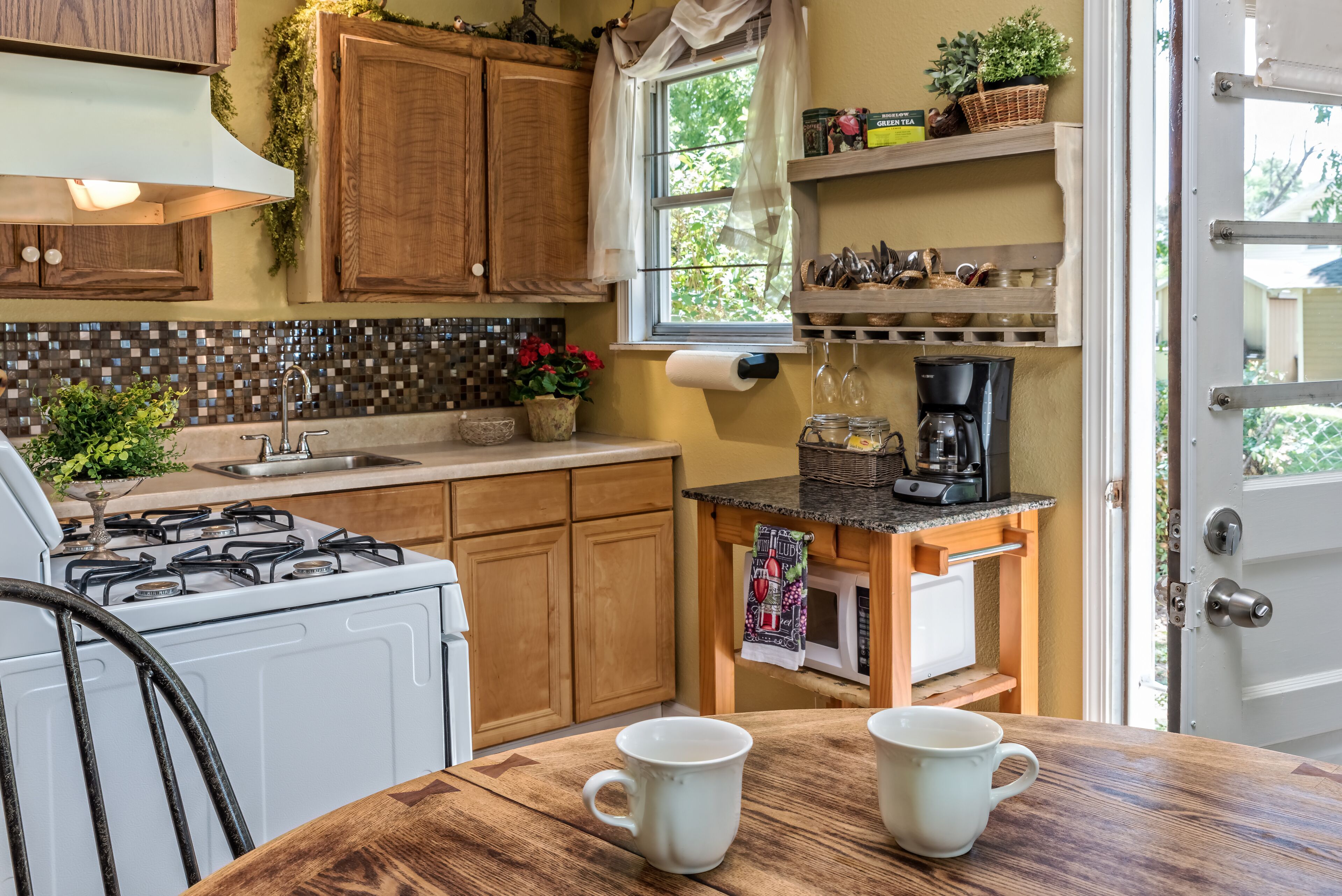 Una cocina con armarios de madera, una estufa a gas, un lavabo y una cafetera. Hay dos tazas blancas sobre la mesa.