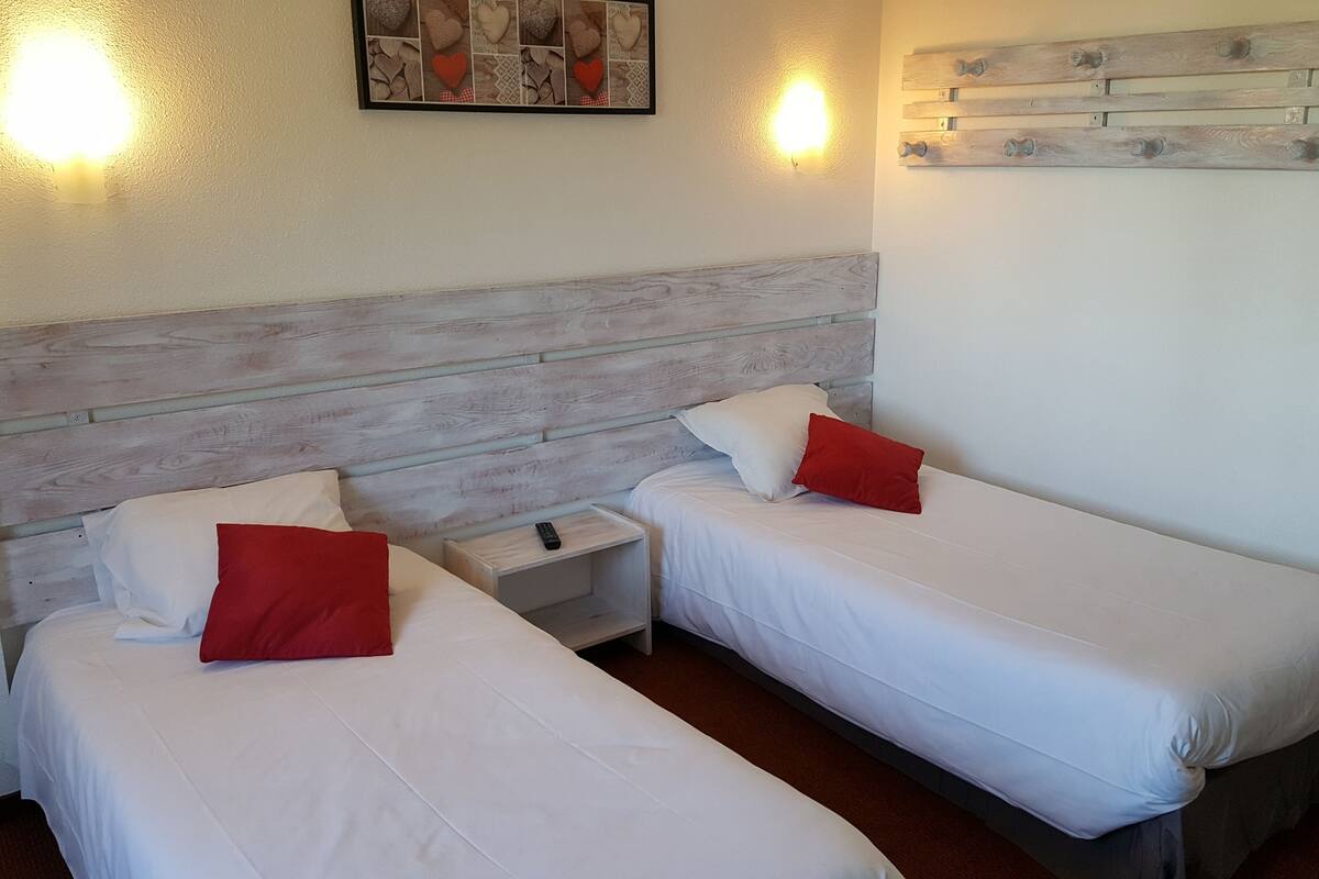 Une chambre d’hôtel avec deux lits simples, chacun doté de draps blancs et d’oreillers rouges. On y trouve une tête de lit en bois, une table de chevet avec une télécommande et un luminaire fixé au mur.