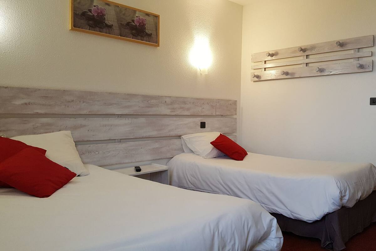 Une chambre d’hôtel avec deux lits, chacun doté de draps blancs et d’oreillers rouges. On y trouve une tête de lit en bois, une lampe fixée au mur et une étagère en bois avec des crochets.