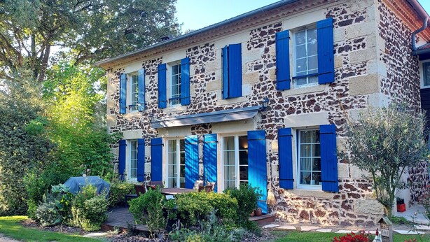 Un edificio de piedra de dos plantas con contraventanas azules, un porche cubierto y un jardín con flores rojas.