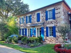 A two-story stone building with blue shutters, a covered patio, and a garden with red flowers. - Chambres d'hôtes L'Airial (Liposthey)