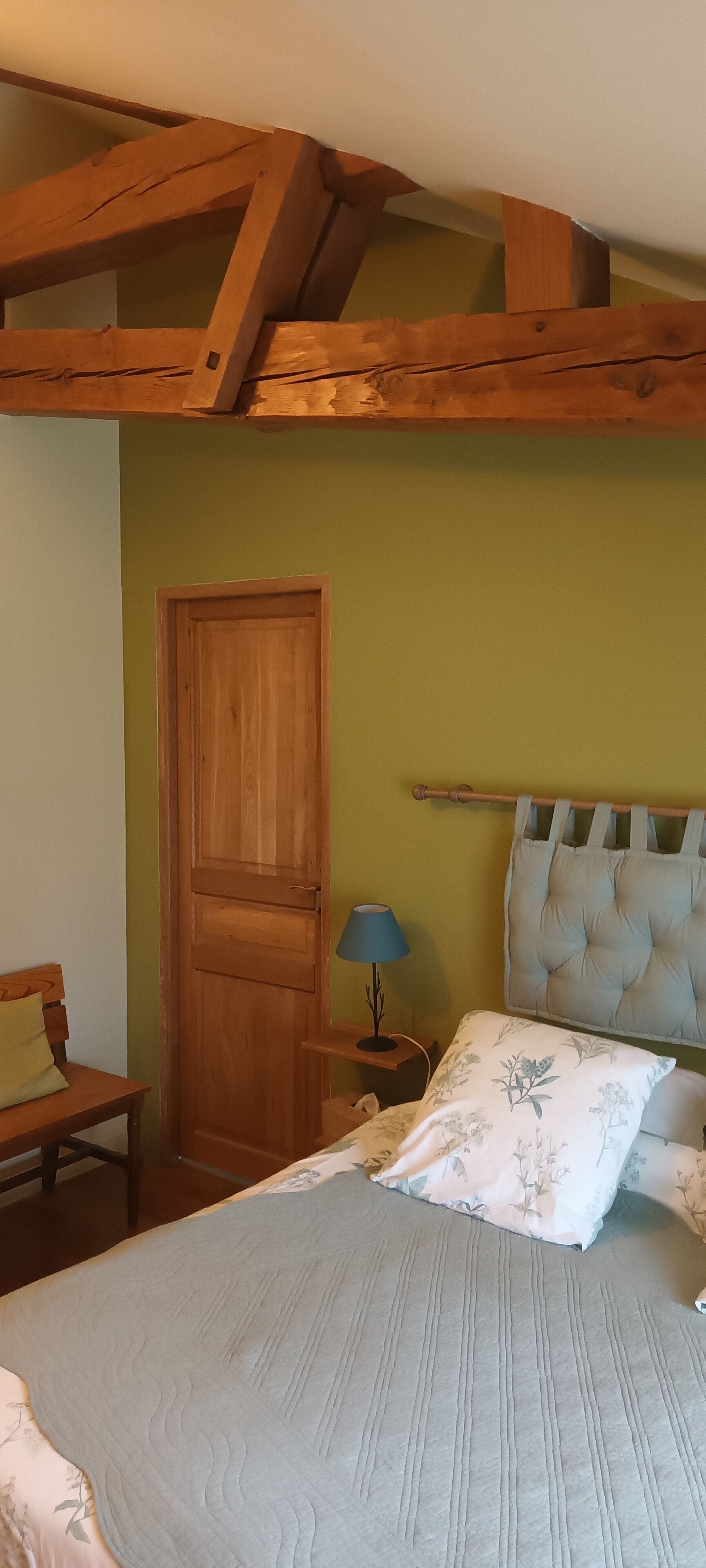 Una camera da letto con soffitto in legno, un letto con cuscino floreale, un comodino con una lampada e una porta in legno.