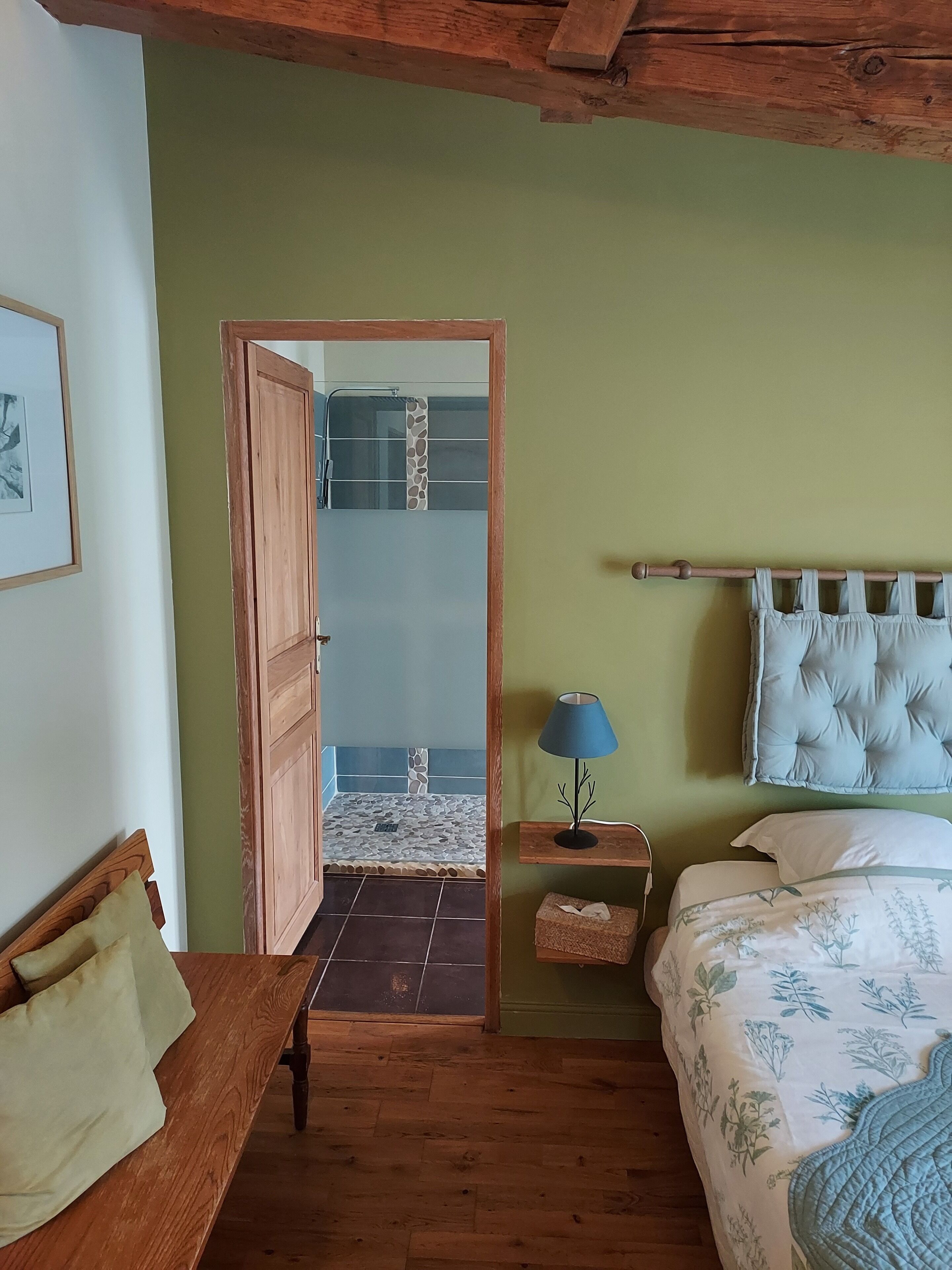 A bedroom with a wooden bed, a nightstand with a lamp, a bench, and a door leading to a bathroom with a tiled floor.