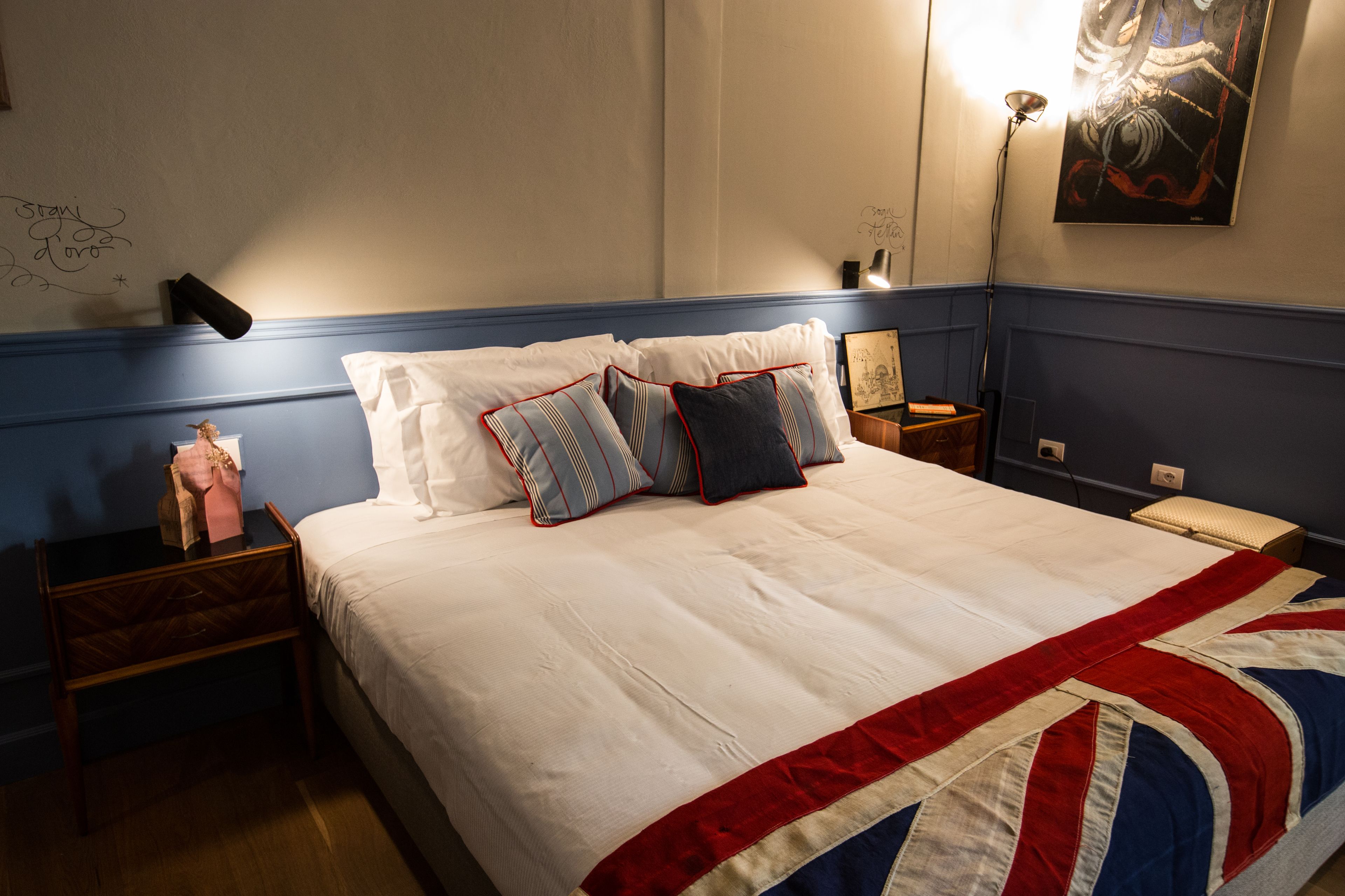 Una cama bien hecha con una manta blanca y roja, una mesita de noche de madera con una lámpara y una pequeña mesa con un objeto decorativo.