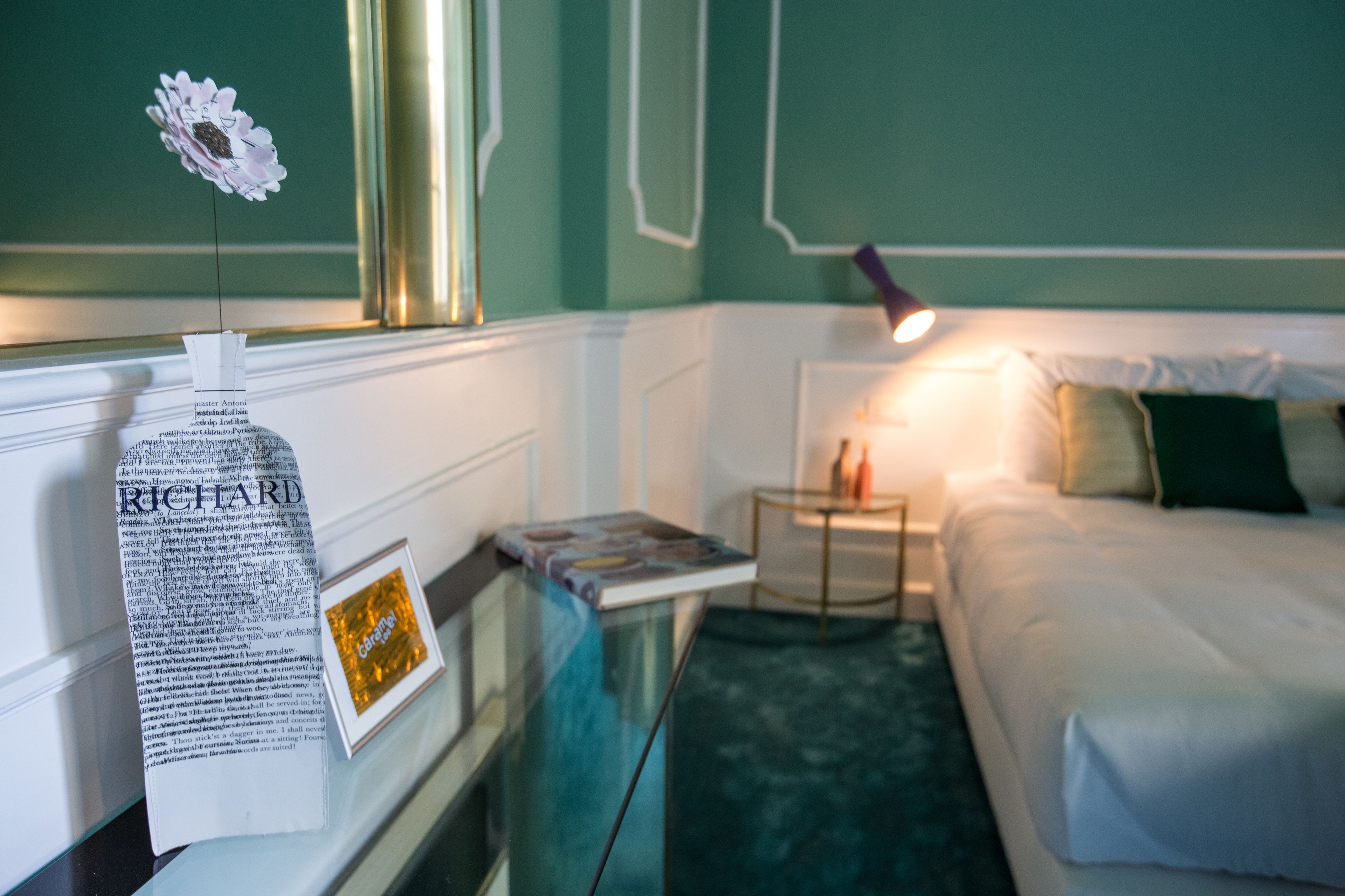 A hotel room with a bed, a bedside table with a lamp, a framed picture, and a decorative flower.