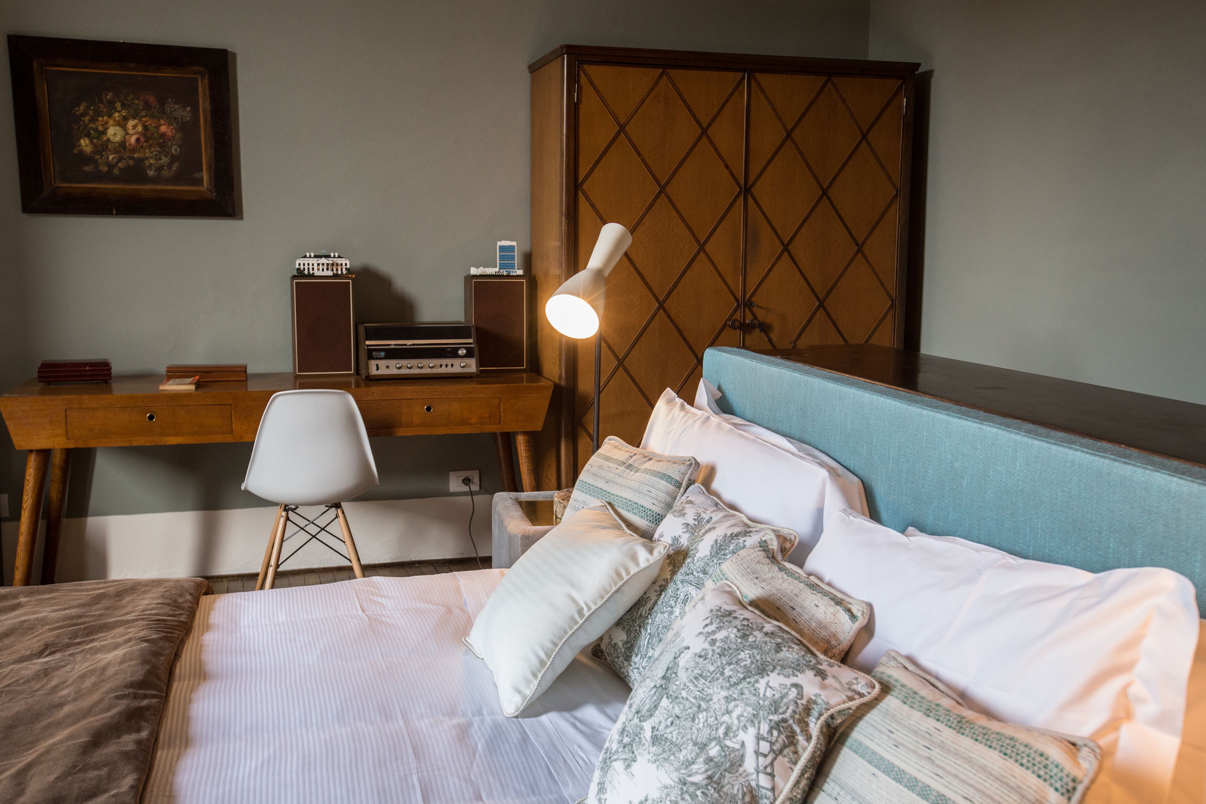 A neatly arranged bedroom with a bed, pillows, a desk with a lamp, and a wardrobe.