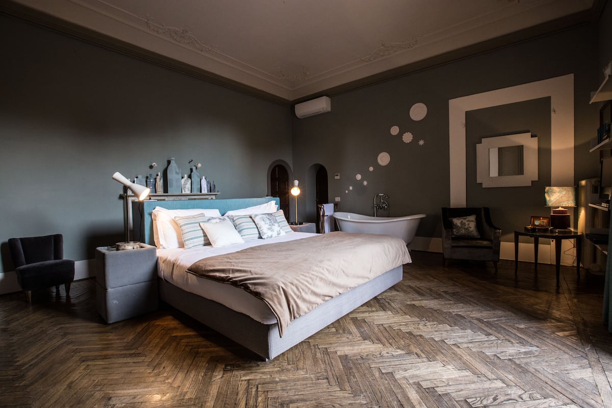 A modern bedroom with a large bed, a nightstand, a chair, a vanity with a mirror, and a bathtub.