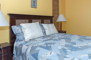 A bed with a patterned bedspread, two white pillows, a wooden headboard, and a nightstand with a lamp. - Sea Shell Beach Studio At Montego Bay Club Resort (Montego Bay)