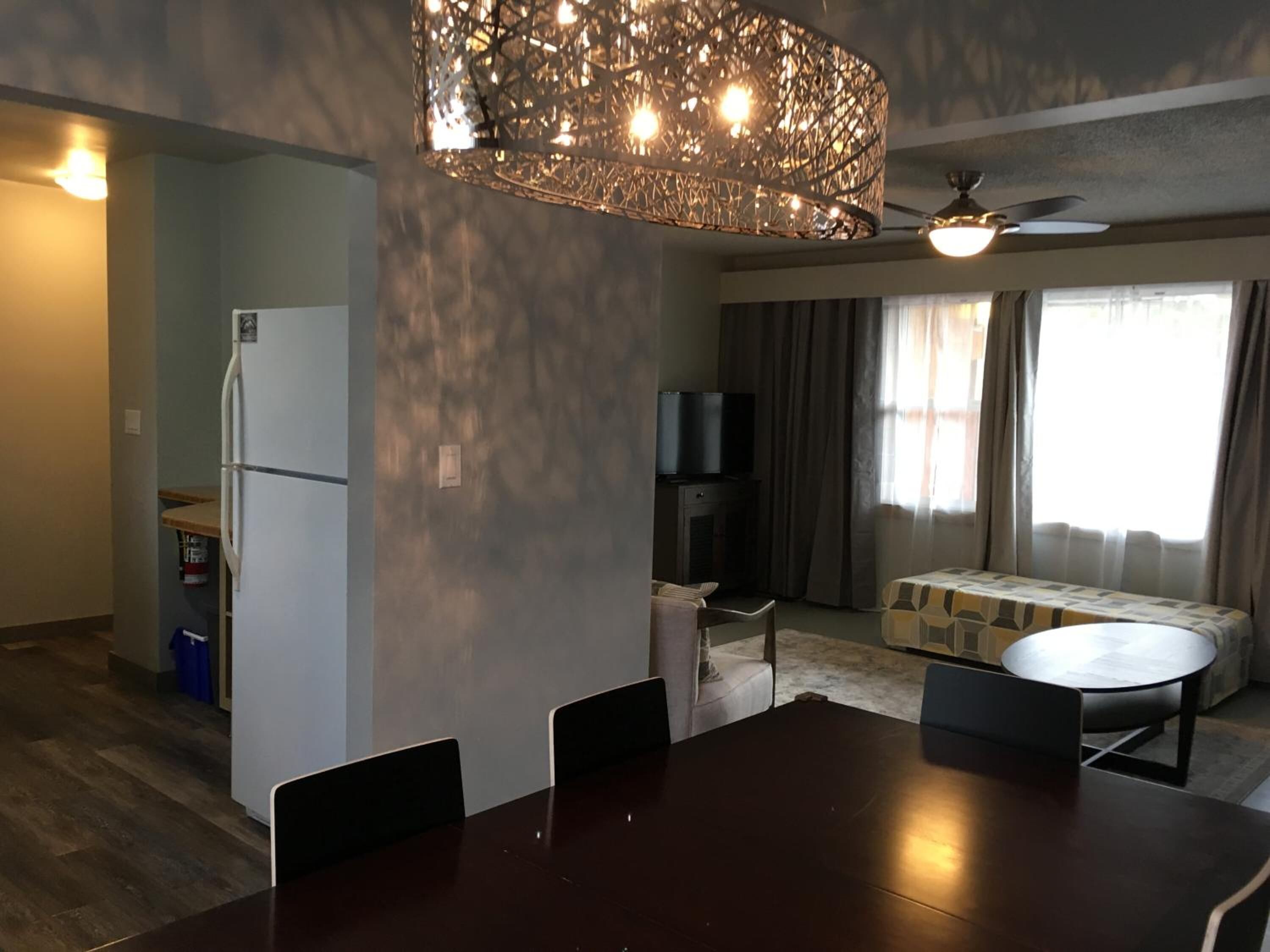 A modern living room with a dark wooden dining table, two chairs, a sofa, and a television. A decorative light fixture hangs from the ceiling.