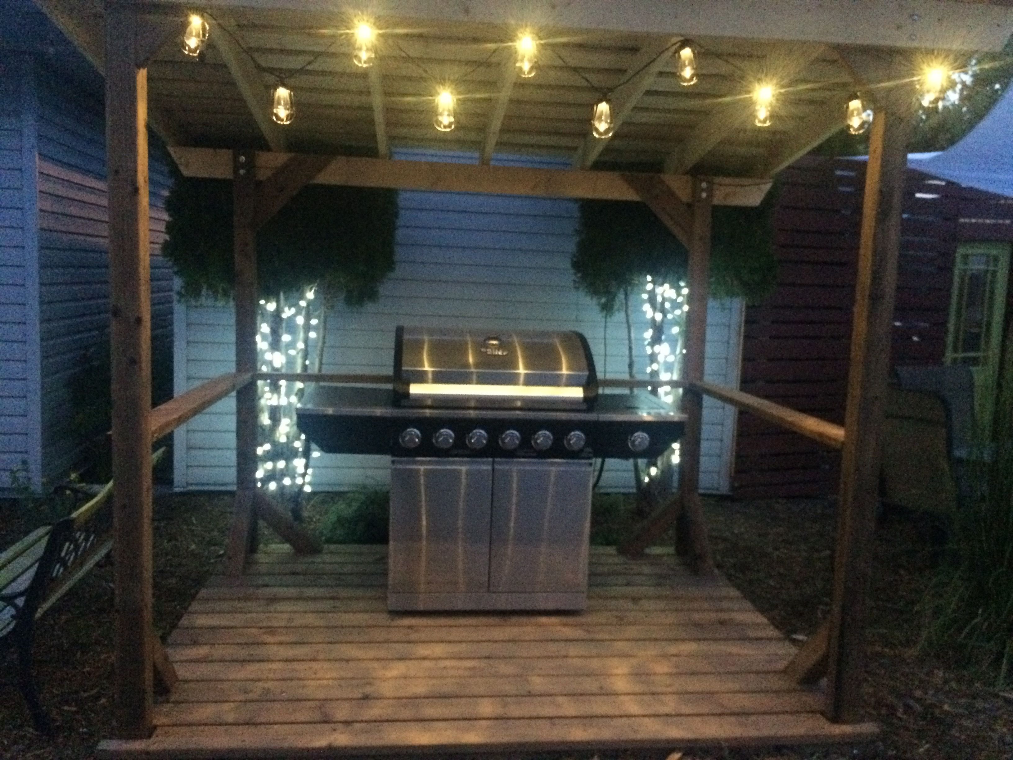 Een buitenbarbecueplaats met een houten pergola, lichtjes en een barbecue met vier branders.