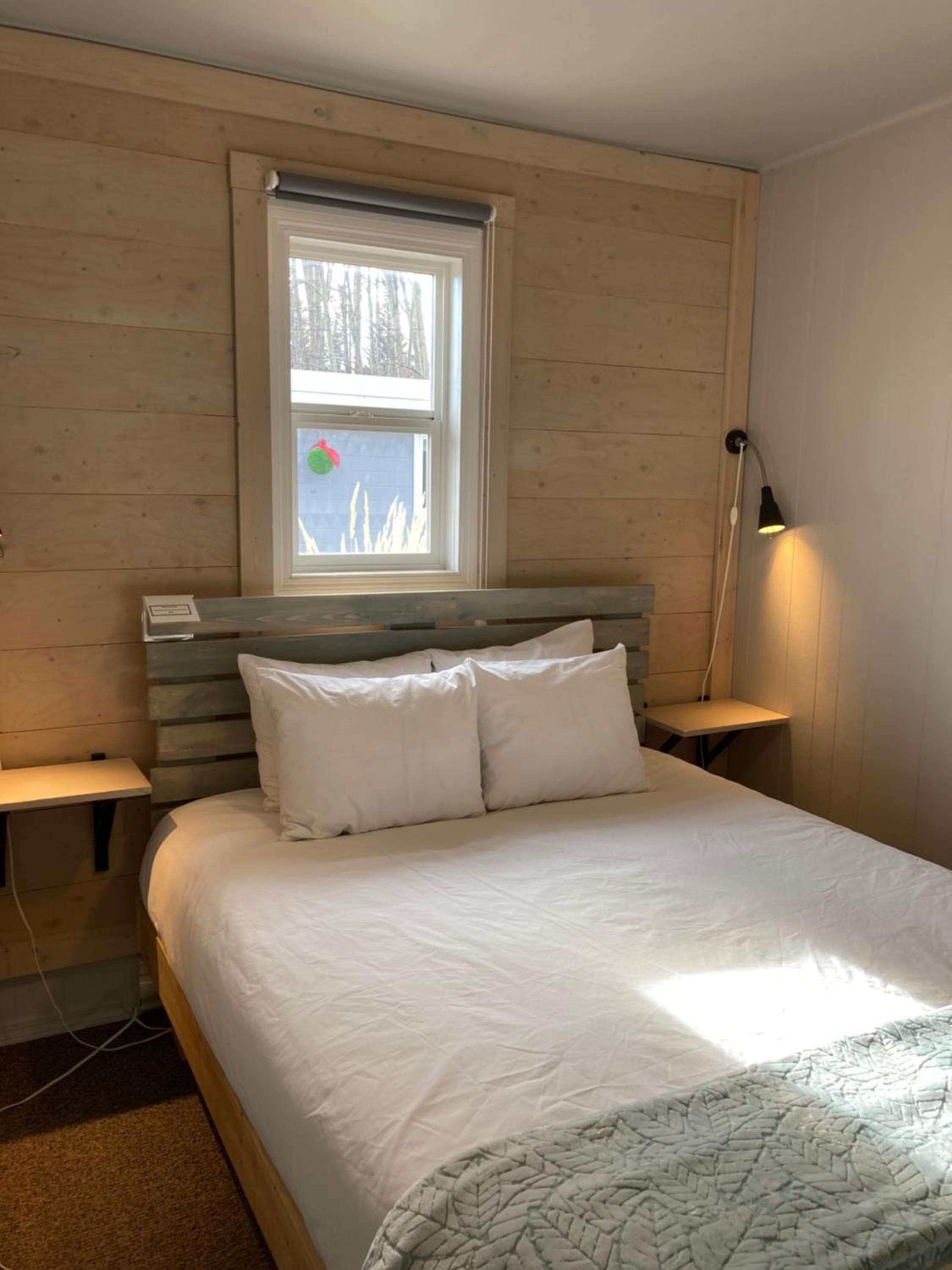A bedroom with a wooden headboard, a large bed with white bedding, a window with a view of trees, and a bedside lamp.