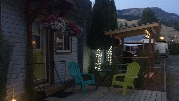 A wooden deck with two green chairs and a table, a porch with a flower arrangement, and string lights. - Pinewood Inn (Golden)
