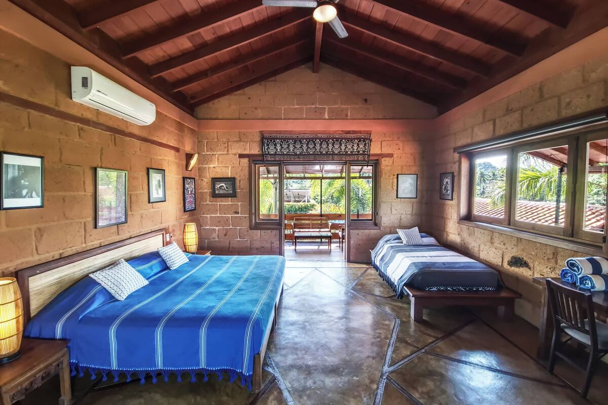 A room with a high ceiling, brick walls, and a large window. There are two beds with blue bedding, a bench, and a chair.