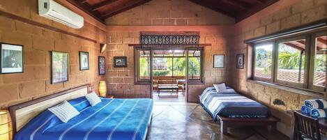 A room with a high ceiling, brick walls, and a large window. There are two beds with blue bedding, a bench, and a chair.