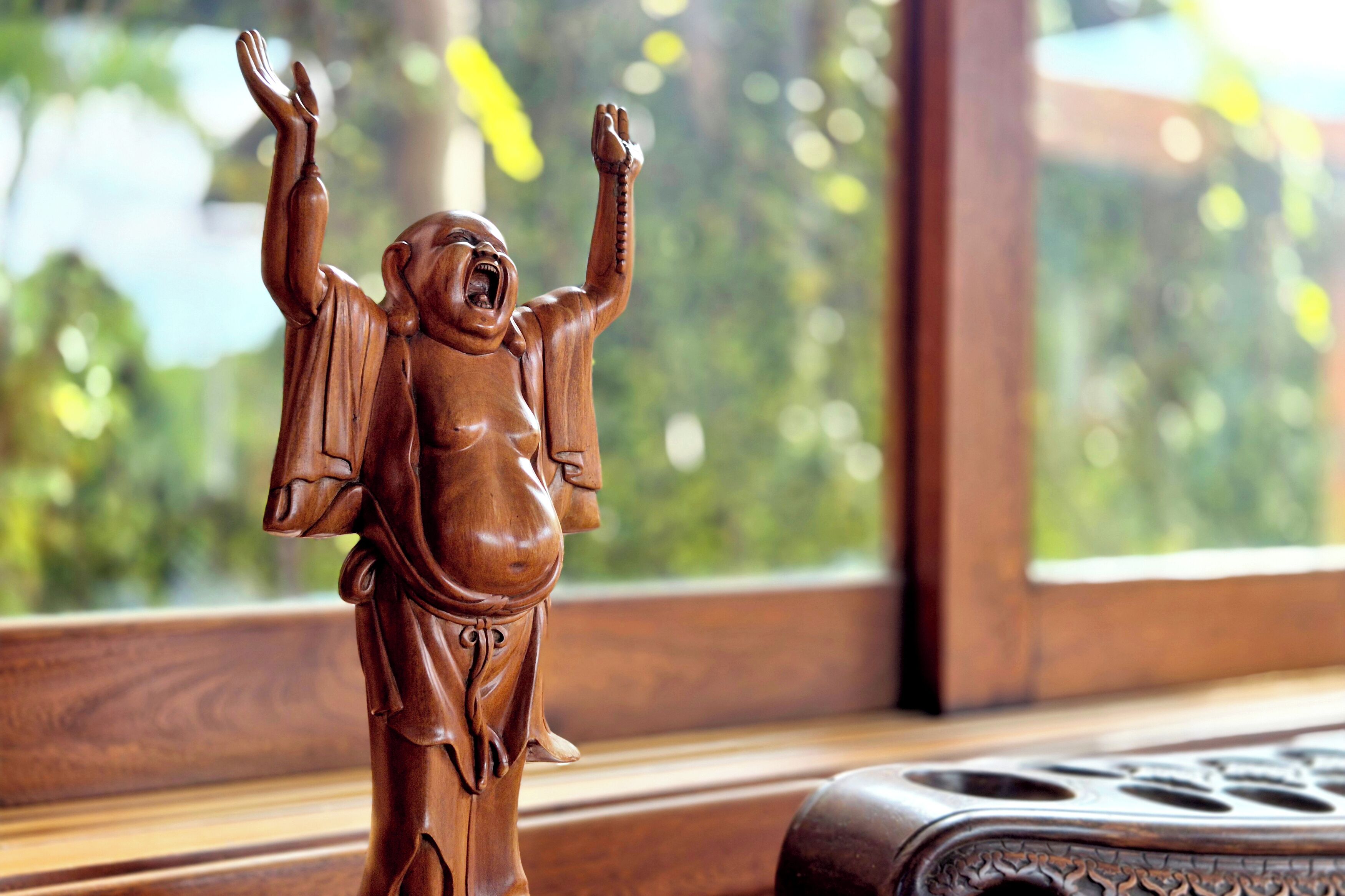 A wooden statue with raised arms, placed on a wooden surface near a window with a blurred green background.