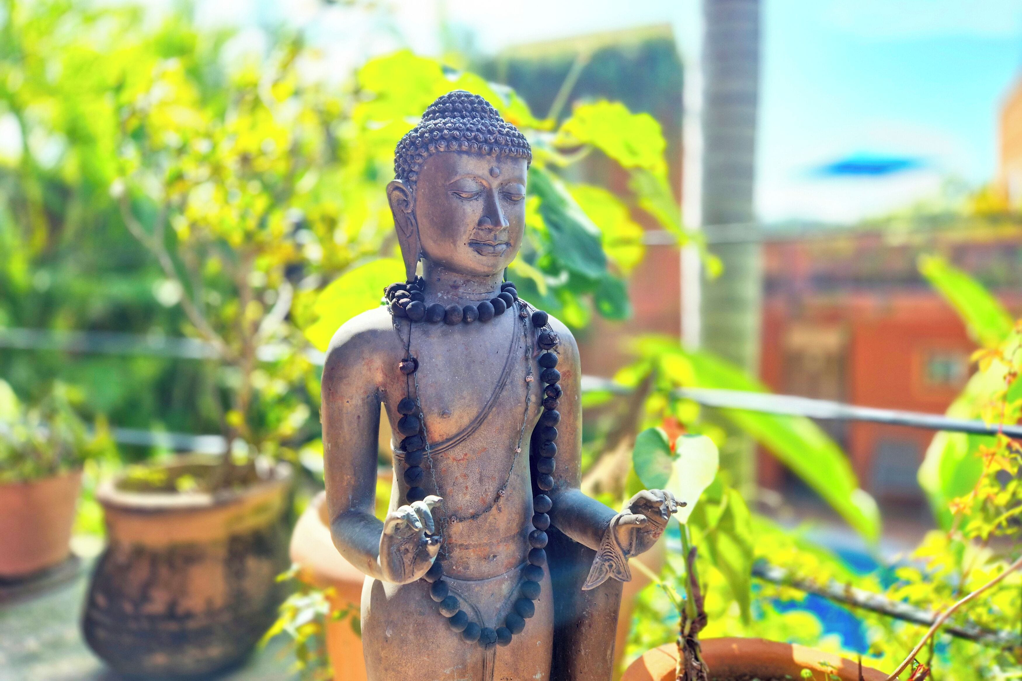 Una estatua de una figura con cabeza redondeada y cuerpo alargado, adornada con un collar de cuentas y un paño ceñido a la cintura, frente a un fondo de exuberante vegetación y un edificio.