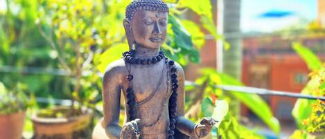 A statue of a figure with a rounded head and elongated body, adorned with a beaded necklace and a draped cloth around the waist, set against a backdrop of lush greenery and a building.