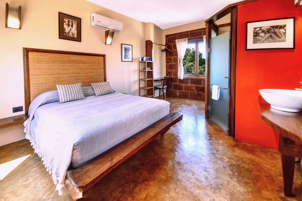 A bedroom with a wooden bed, a wooden desk, a red accent wall, and a bathroom with a white sink.