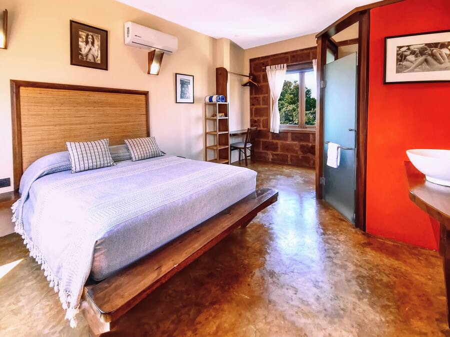 Une chambre avec un lit en bois, un bureau en bois, un mur d’accent rouge et une salle de bain avec un lavabo blanc.