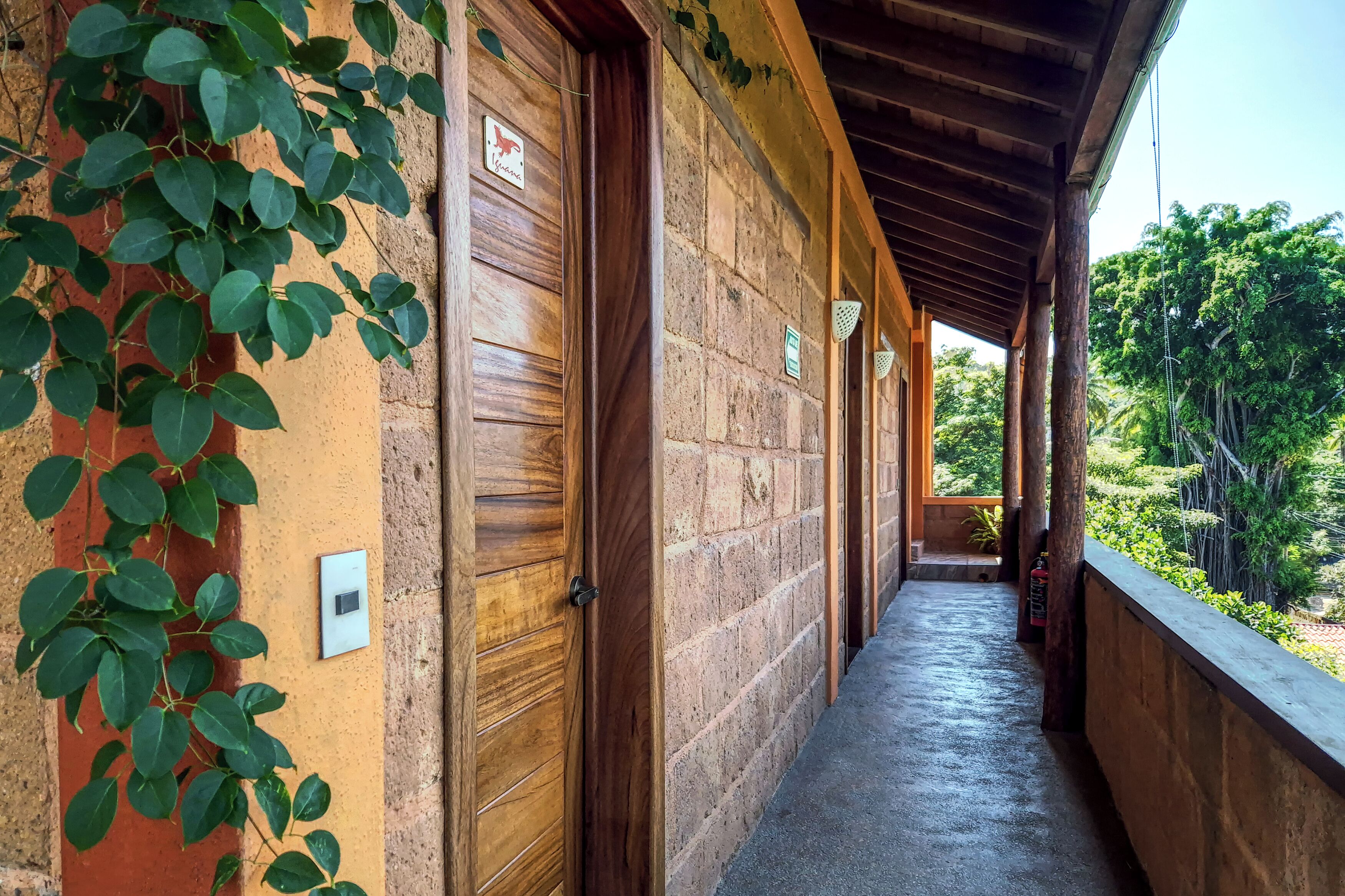 A covered corridor with wooden doors and a brick wall, leading to a balcony with a view of greenery.