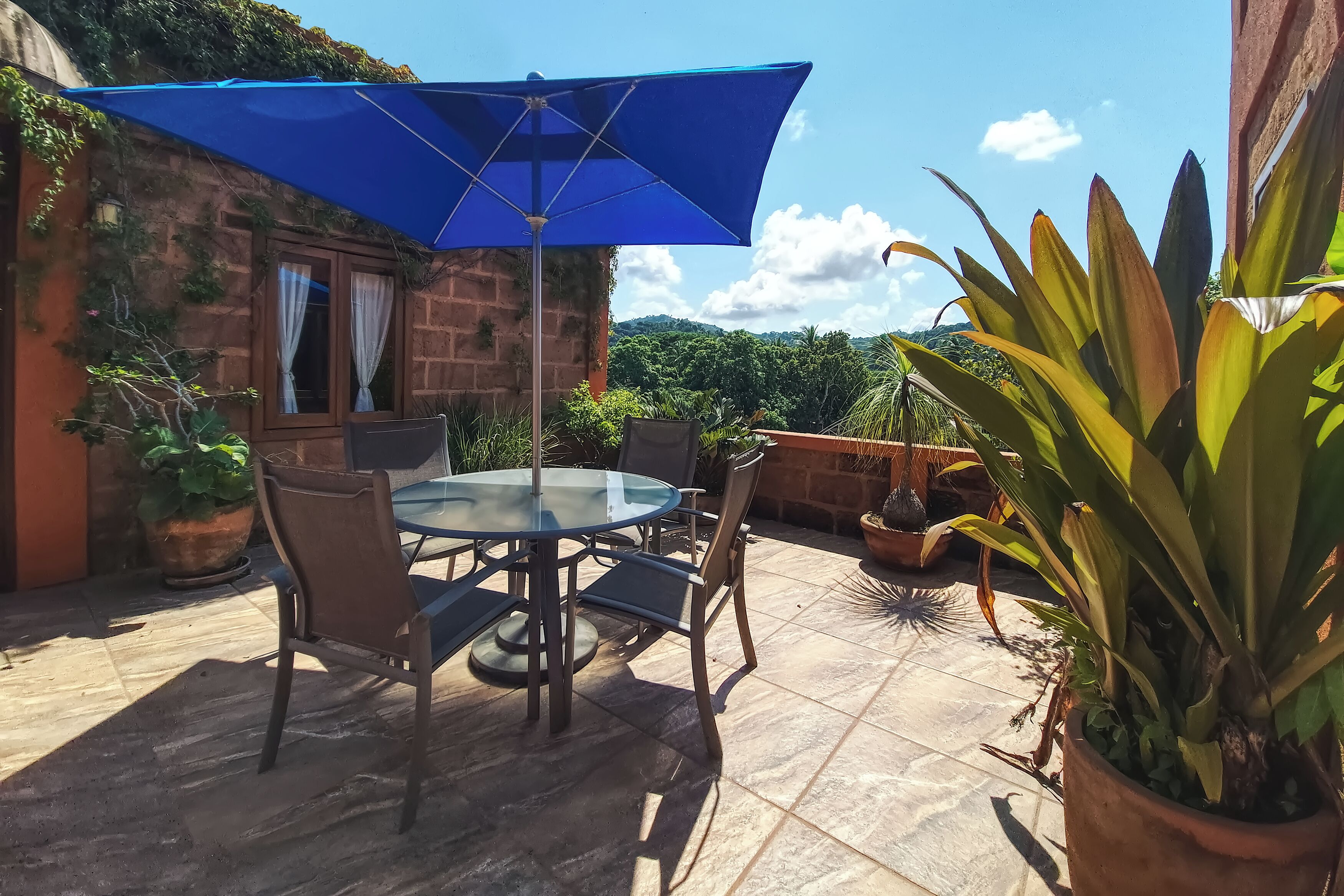 A patio with a glass table and chairs, a blue umbrella, and potted plants.