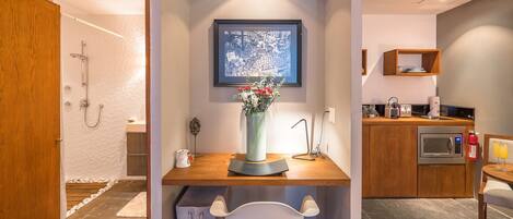 Habitación compacta y moderna con escritorio de madera, una silla blanca, una obra de arte enmarcada en la pared y una pequeña kitchenette con microondas y tostadora.