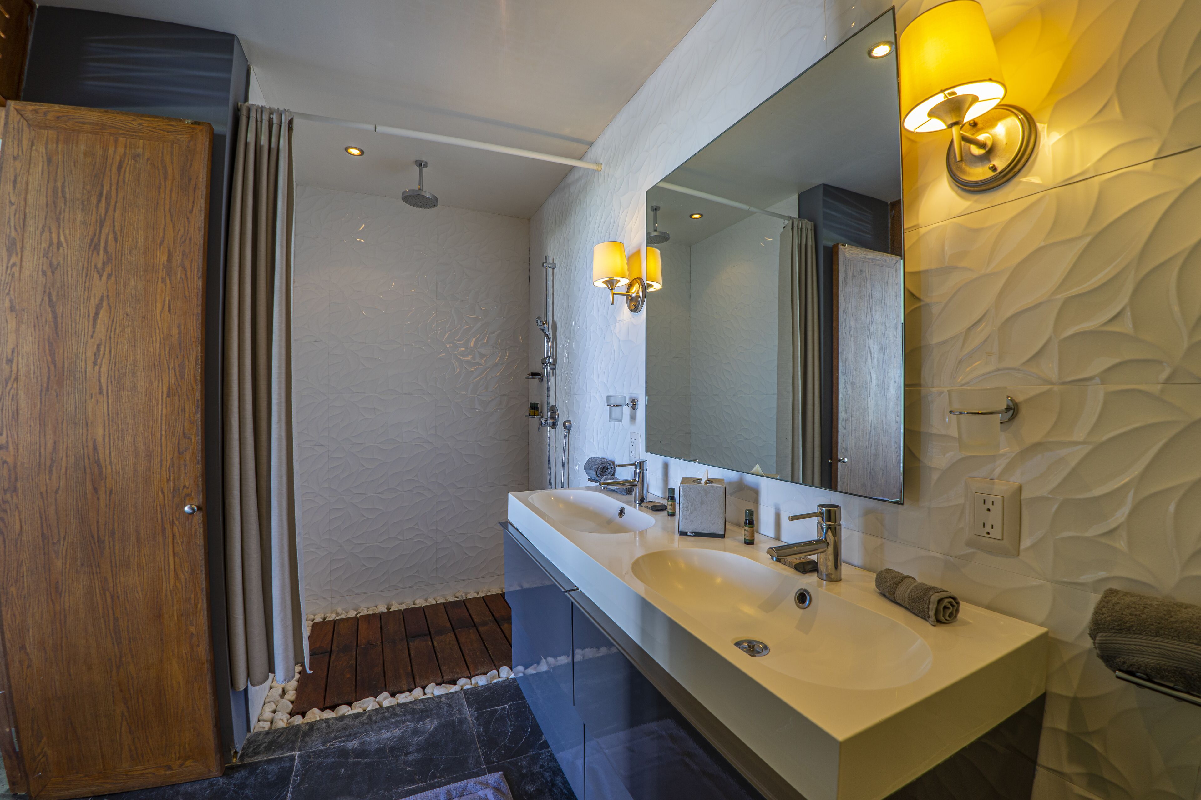 A modern bathroom with a large mirror, two sinks, a shower, and a wooden door.