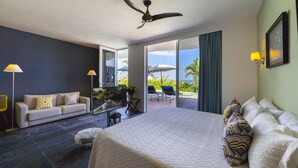 A hotel room with a large bed, a sofa, a TV, and a balcony with chairs and an umbrella. - Suites at Sapphire Ocean Club (Puerto Vallarta)