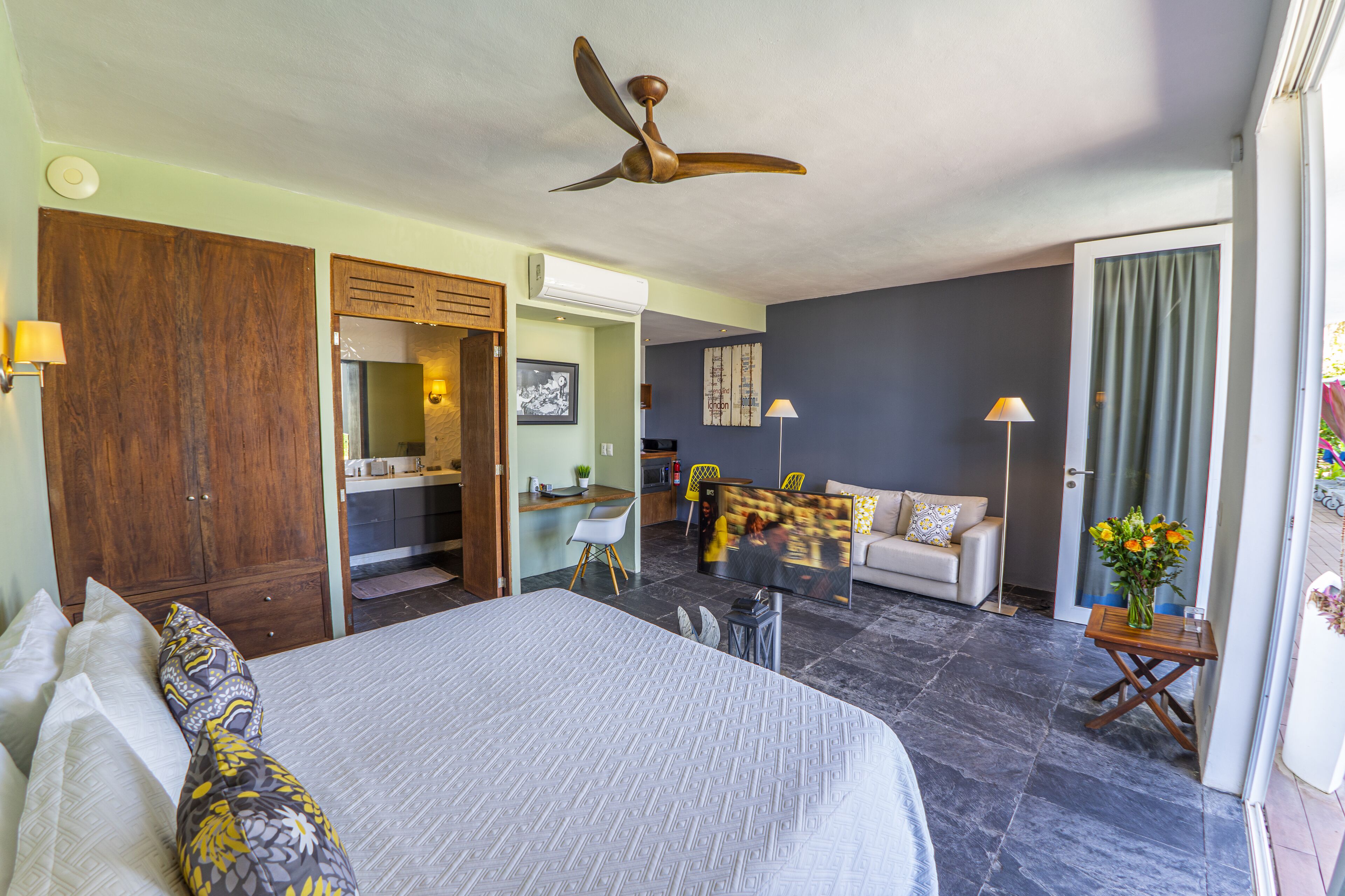 Une chambre d’hôtel avec un grand lit, un ventilateur de plafond, une télévision, un canapé et une petite table agrémentée de fleurs.