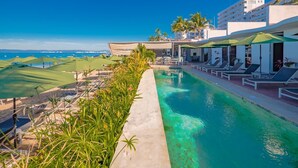 A poolside area with lounge chairs and umbrellas, a view of the ocean, and a building with balconies. - Suites at Sapphire Ocean Club (Puerto Vallarta)