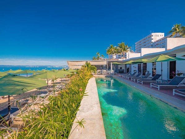 A poolside area with lounge chairs and umbrellas, a view of the ocean, and a building with balconies.