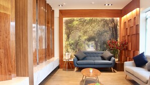 A modern living room with a blue sofa, a white sofa, a wooden coffee table, and a view of a forest path through a large window. - Habitat Hotel (Lima)